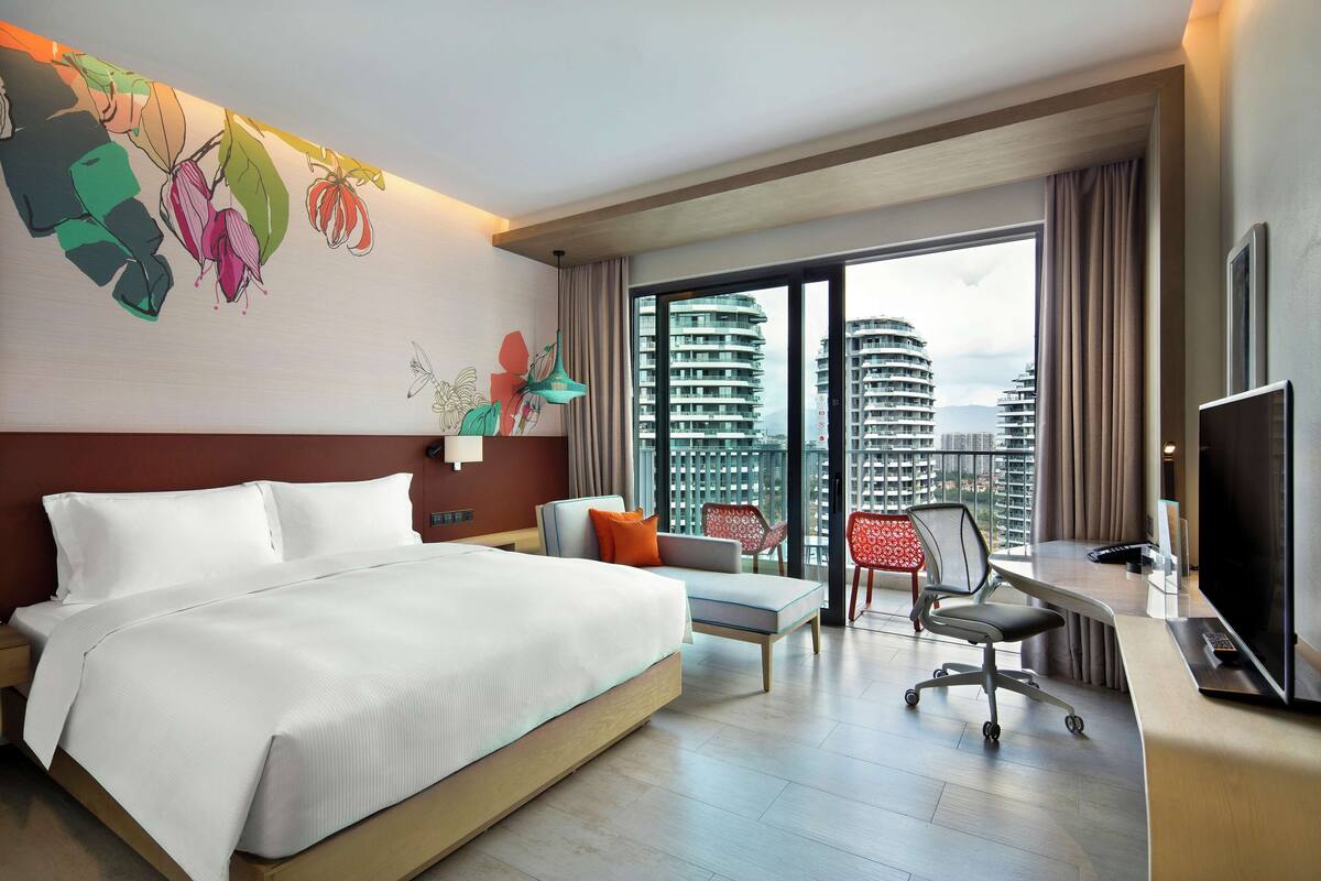 A hotel room with a large bed, a desk with a TV, a chair, and a balcony with a view of high-rise buildings.