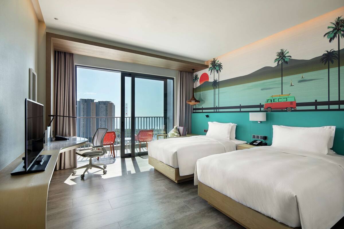 A hotel room with two beds, a desk with a computer, a chair, a TV, and a balcony with a view of the city.