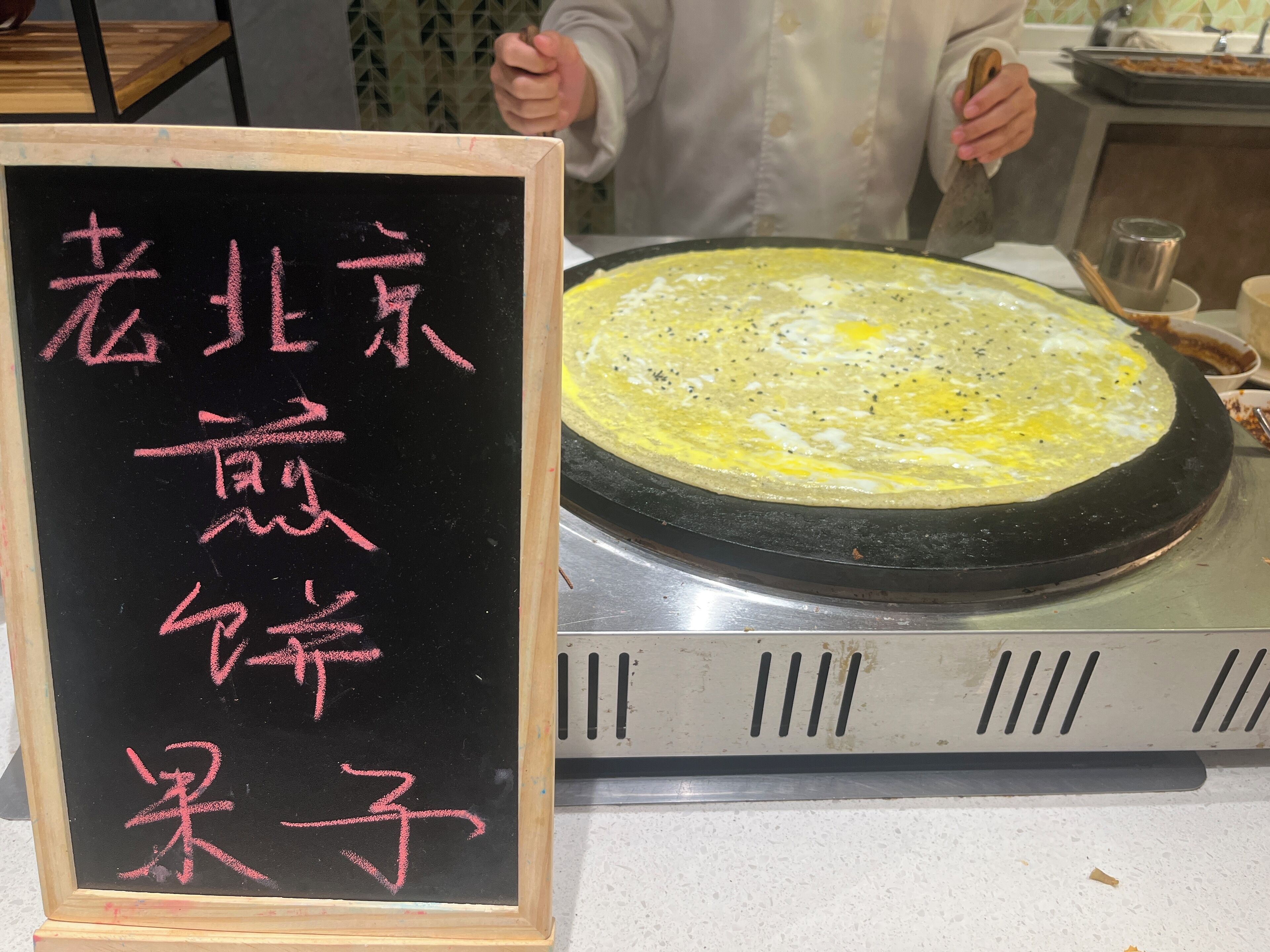 A blackboard with pink text, a large griddle with a pancake, and a person cooking.