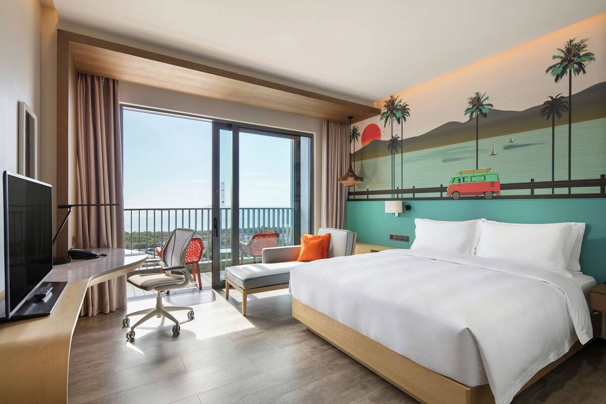 A hotel room with a large bed, a desk with a computer, a chair, and a view of the ocean through large windows.
