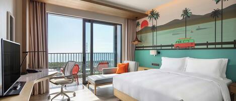 Habitación de hotel con una cama grande, un escritorio con computadora, una silla y vista al océano a través de amplios ventanales.