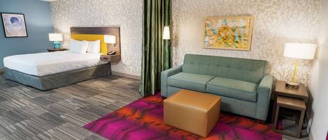 Una habitación de hotel con una cama, un sofá verde, una mesita, una alfombra colorida y un cuadro en la pared.
