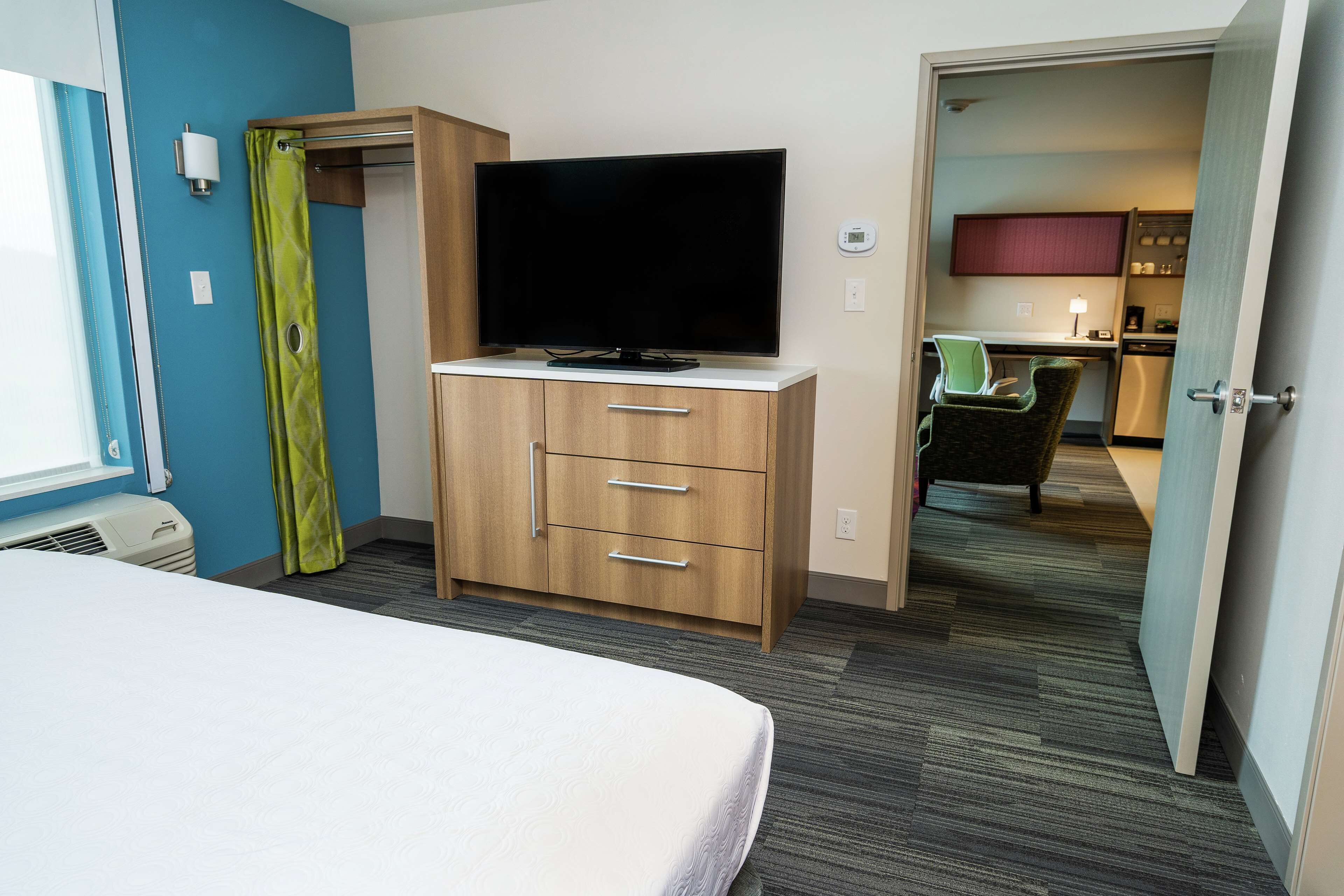 Une chambre d’hôtel avec un lit, un téléviseur posé sur un meuble en bois, un rideau vert et, au fond, une petite cuisine.