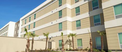 A modern multi-story building with a flat roof, large windows, and a landscaped front yard with palm trees.