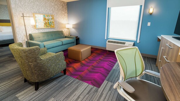 A hotel room with a blue wall, a patterned carpet, a sofa, a chair, a desk, and a window with blinds.