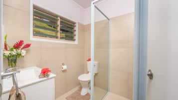 A bathroom with a glass shower enclosure, a toilet, a sink, and a window with blinds.