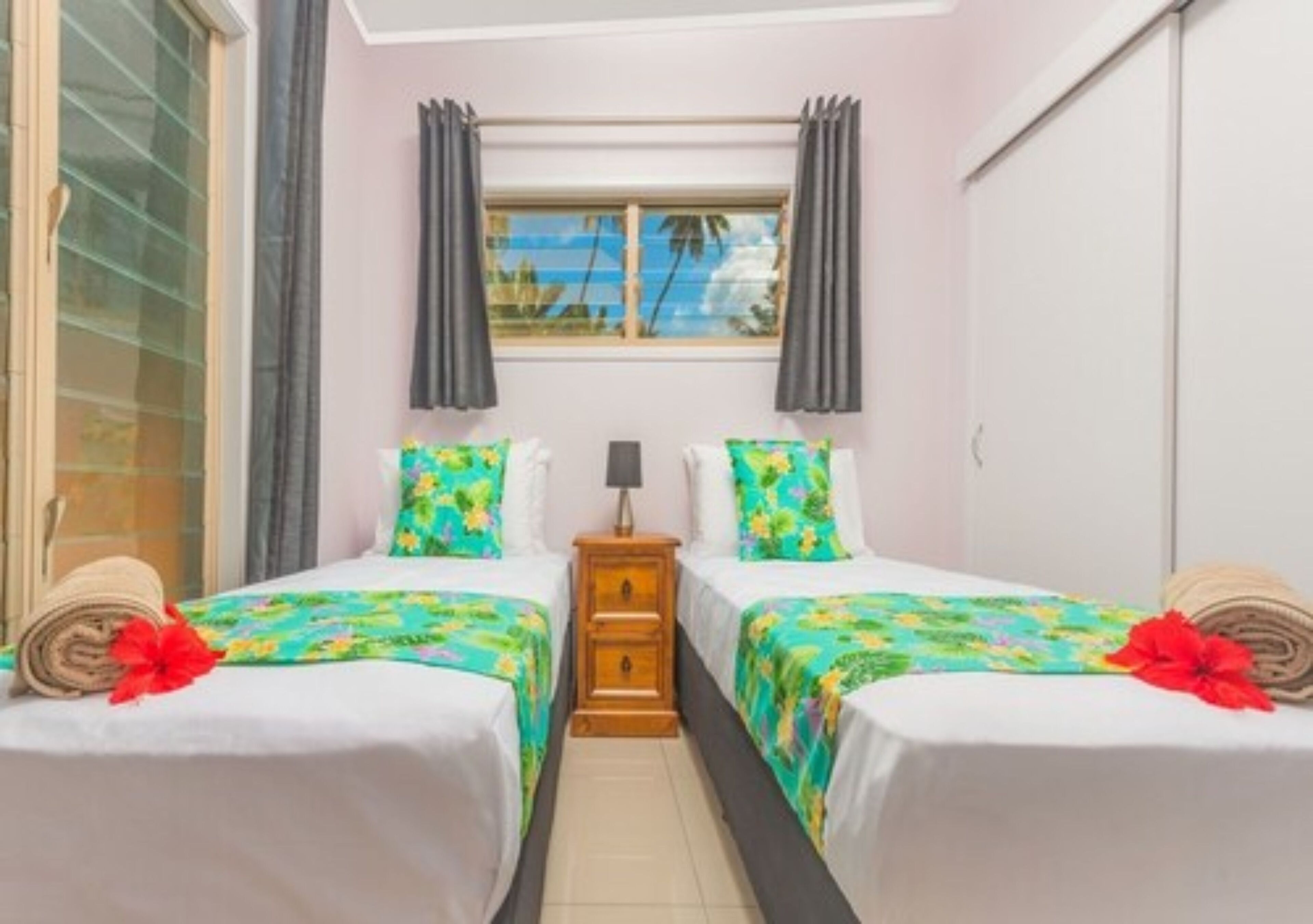 A hotel room with two beds, a window with curtains, a nightstand, and a view of palm trees.