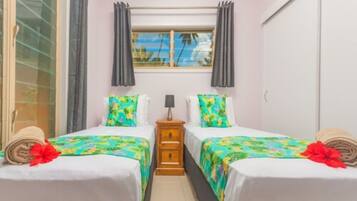 A hotel room with two beds, a window with curtains, a nightstand, and a view of palm trees.