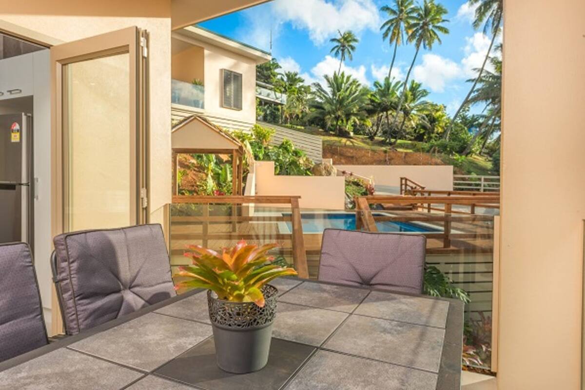 A patio with a table and chairs, a potted plant, and a view of a swimming pool and palm trees.