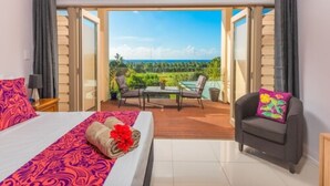 A hotel room with a bed, a small table, a chair, and a view of a garden and ocean through an open door.