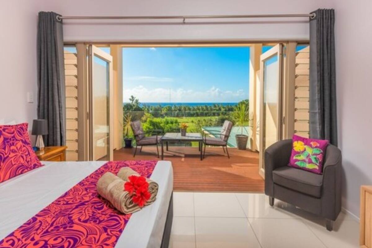 A hotel room with a bed, a small table, a chair, and a view of a garden and ocean through an open door.