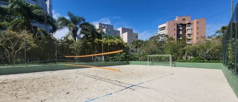 A sandy beach volleyball court with a net, surrounded by greenery and buildings in the background.