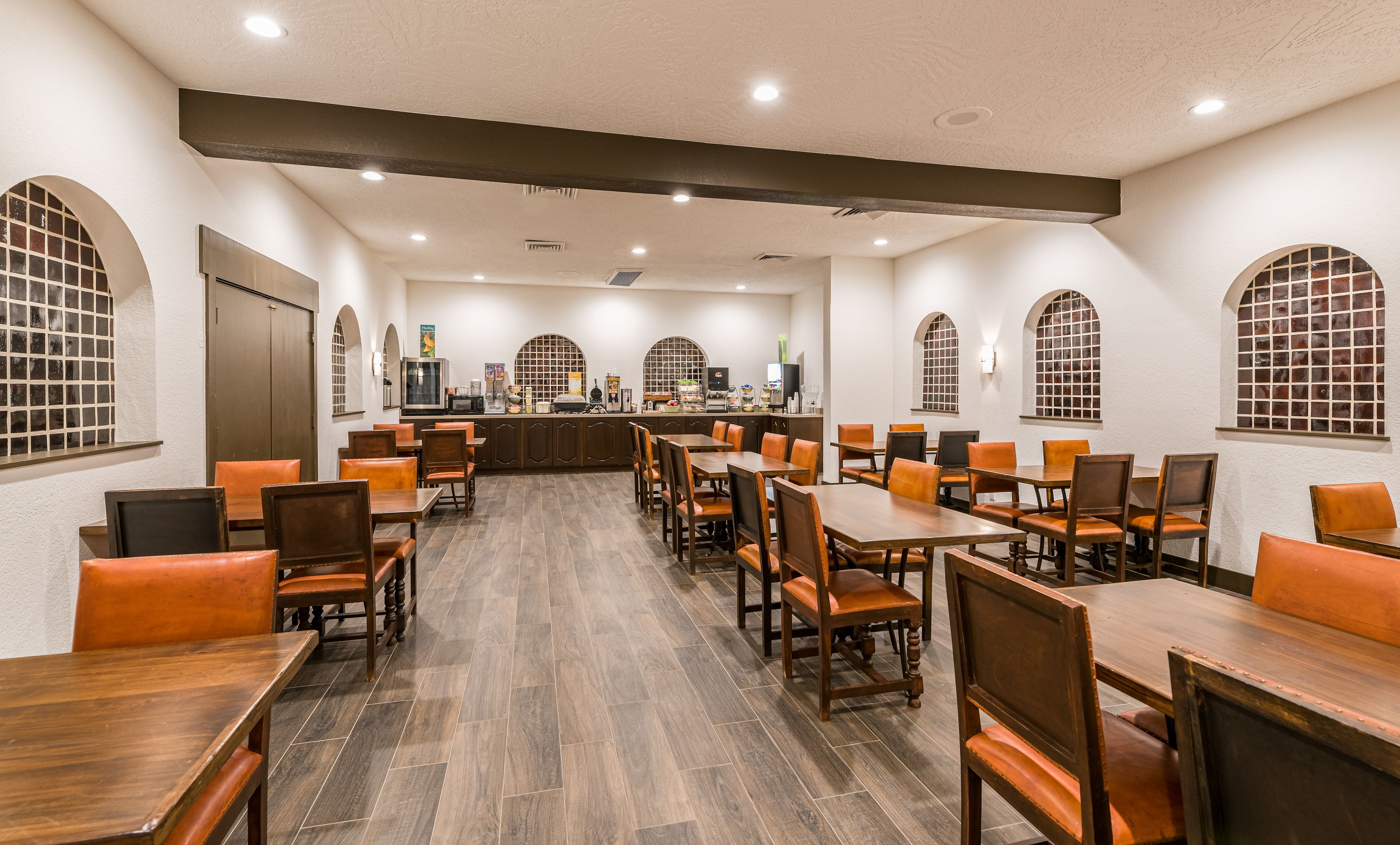 Uma sala de jantar ampla com várias mesas e cadeiras, área de cozinha com eletrodomésticos e janelas em arco.