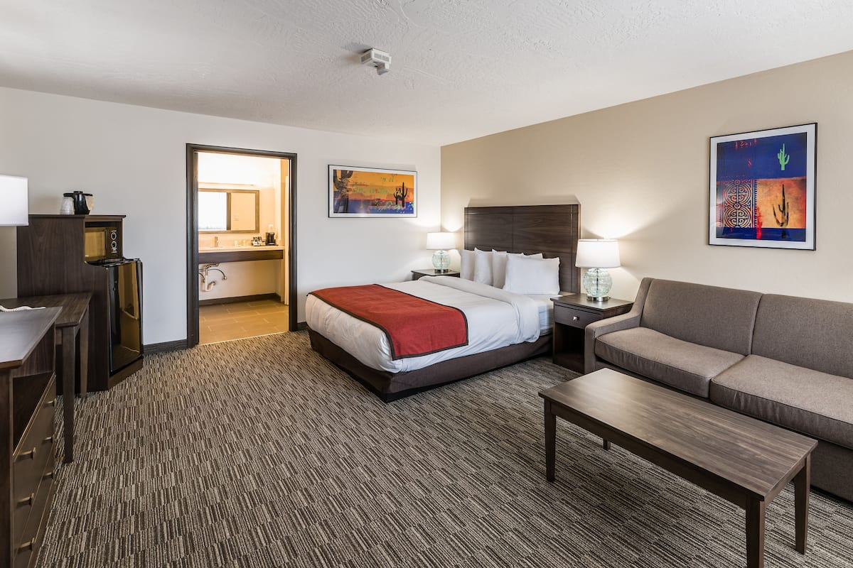 A hotel room with a bed, a sofa, a coffee table, a desk with a lamp, a TV, and a bathroom visible through an open door.