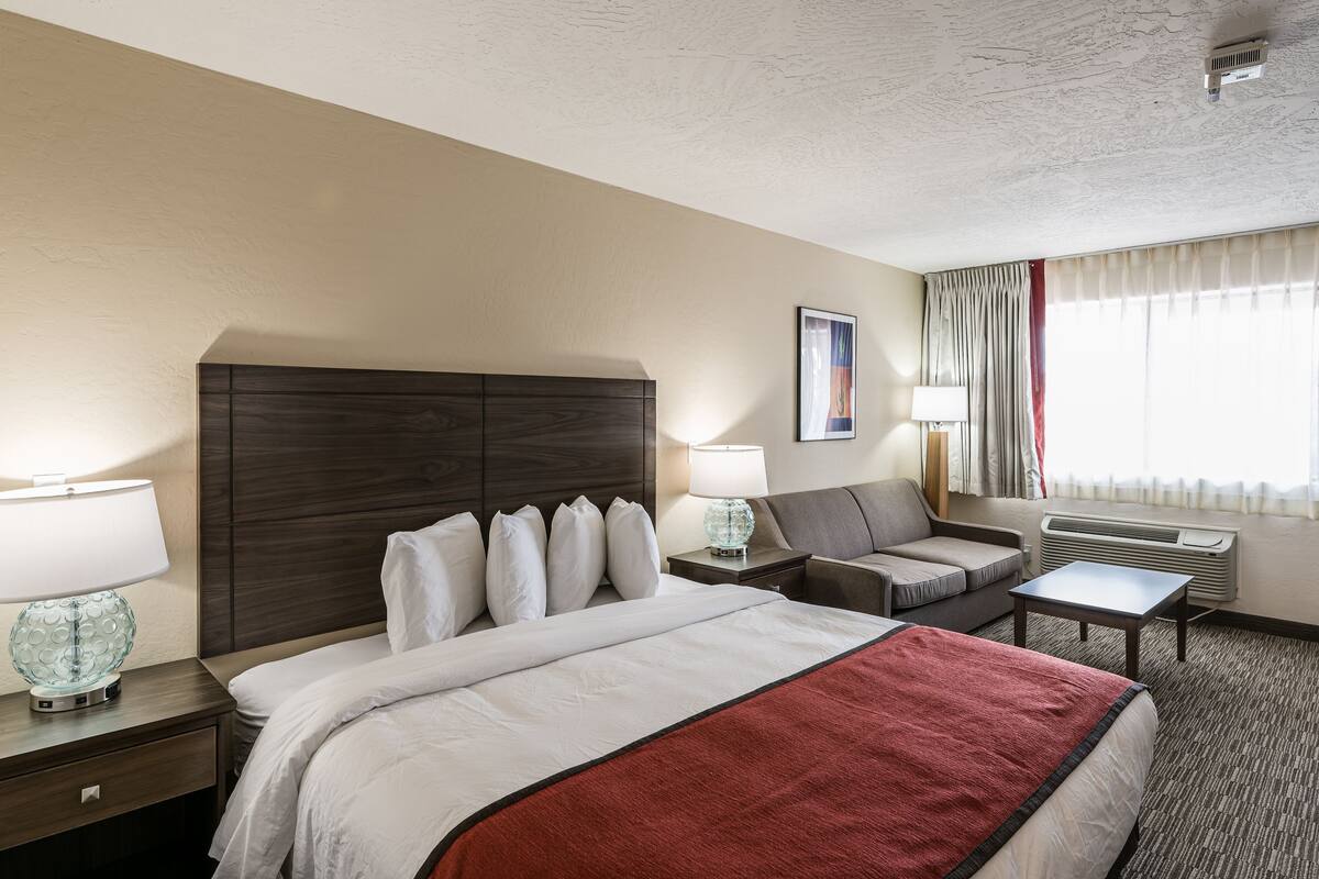A hotel room with a large bed, a desk, a chair, a lamp, and an air conditioning unit.
