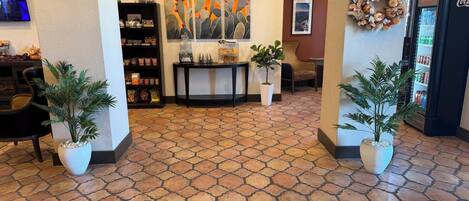 A lobby with a tiled floor, a reception desk, a coffee machine, a wall-mounted TV, a bookshelf, and a refrigerator.