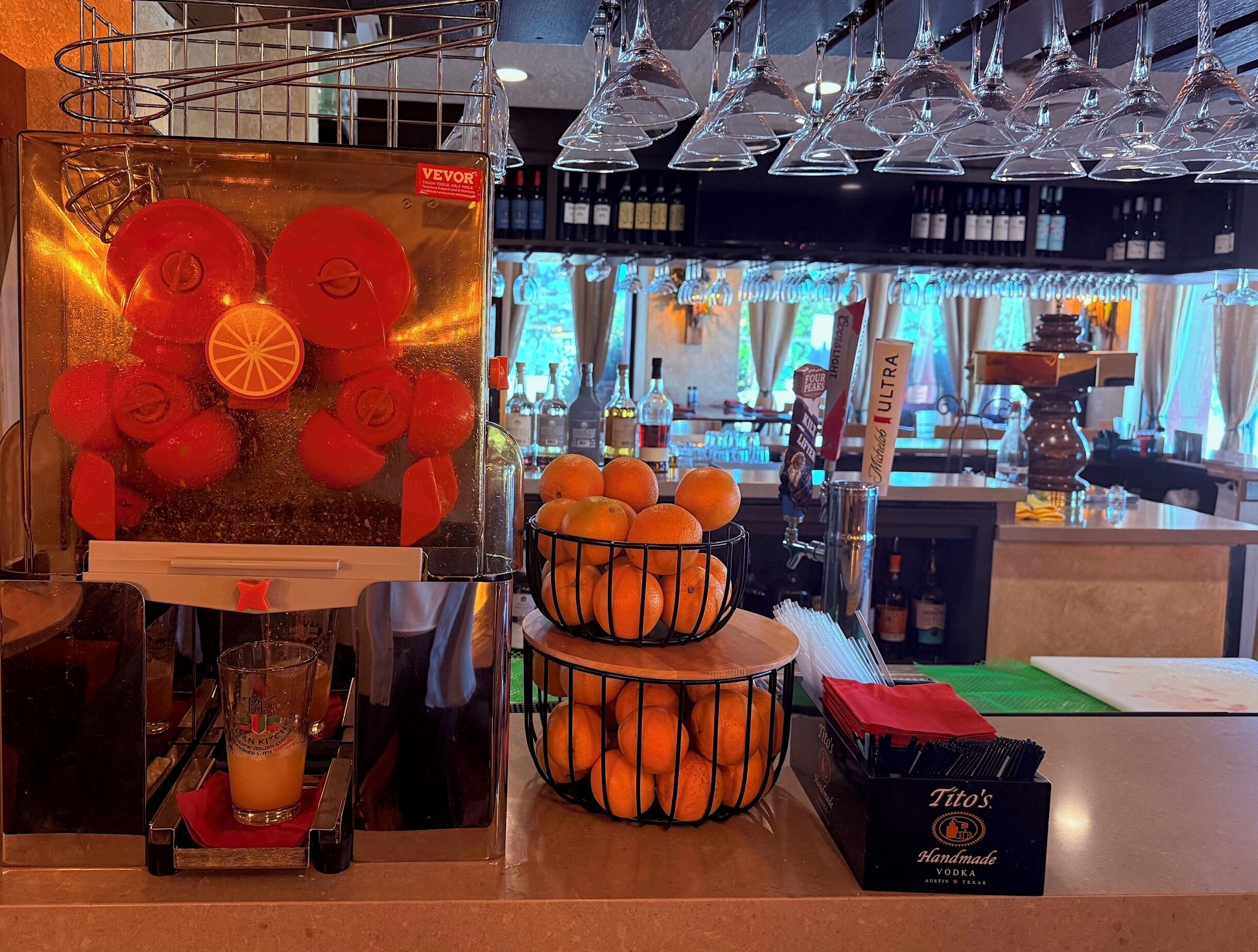 A bar area with a fruit press, a glass of orange juice, a basket of oranges, and a box of vodka.
