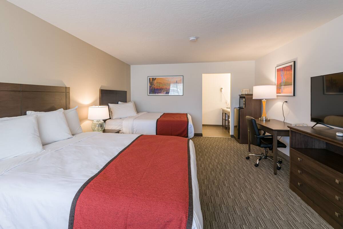 A hotel room with two beds, a desk, a television, and a small kitchenette.