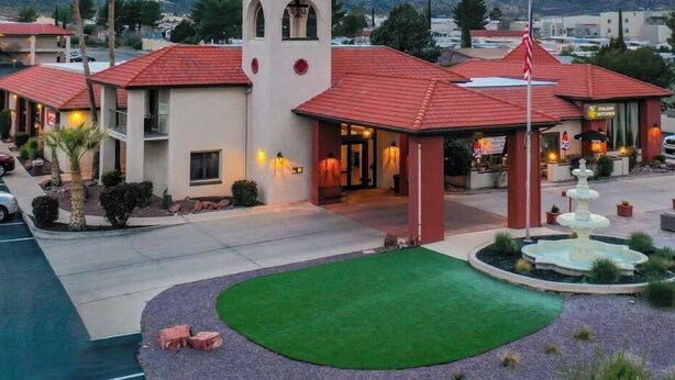 A hotel with a central tower, multiple red-roofed buildings, a landscaped courtyard with a fountain, and a mountainous backdrop. - Lux Verde Hotel (Cottonwood)