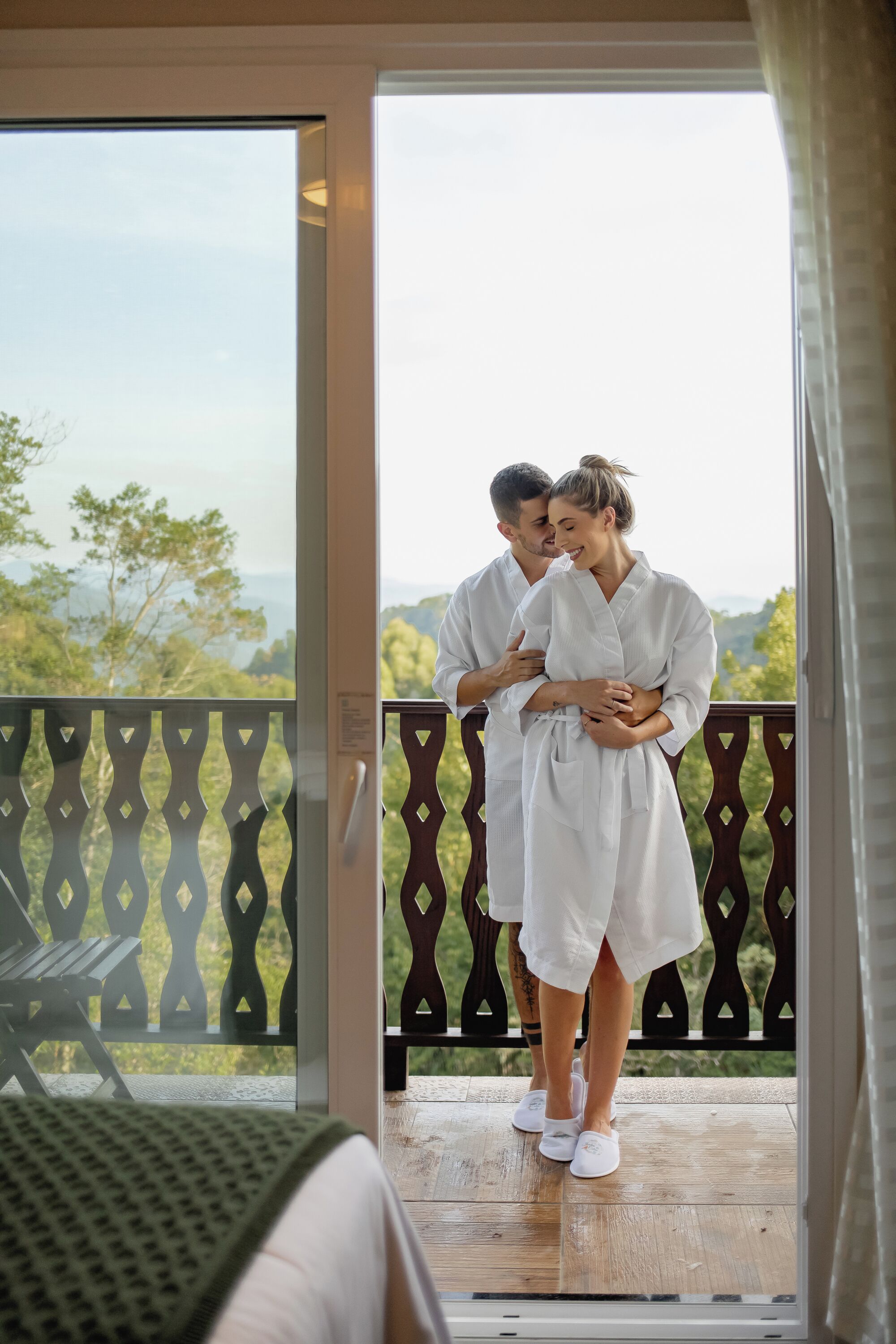 A couple standing on a balcony with a view of greenery and mountains.