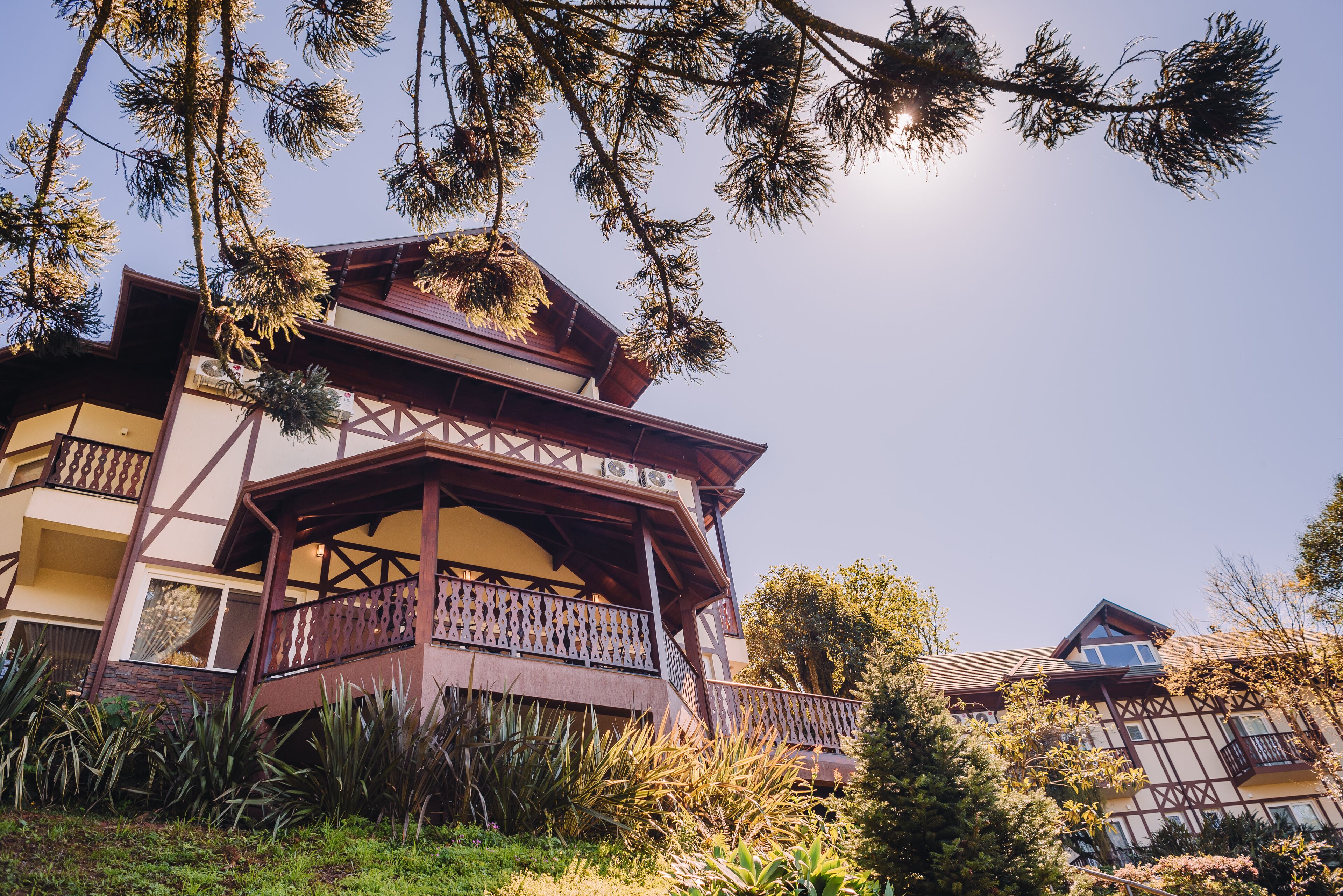 A traditional chalet with a wooden balcony, surrounded by lush greenery and a clear sky.