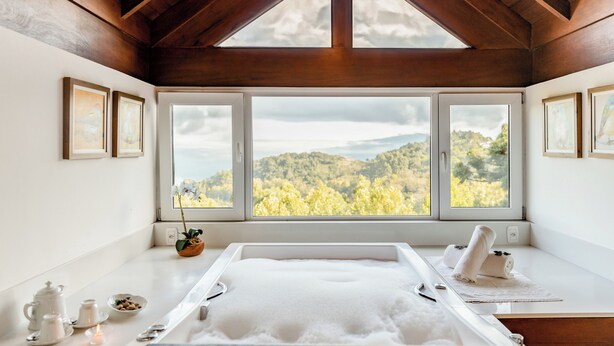 A spa room with a large bathtub, framed artwork, and a view of a forested area through large windows. - Hotel Jardins da Colina (Nova Petropolis)