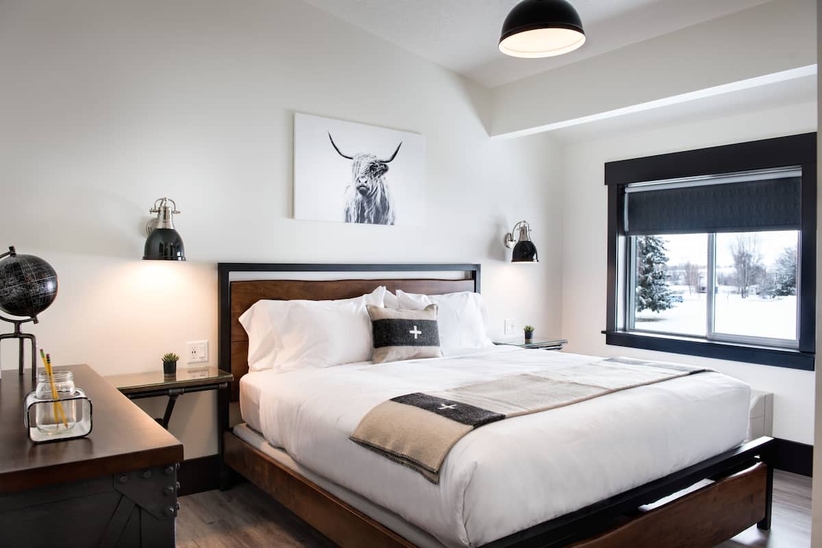 A modern bedroom with a wooden bed, white bedding, a globe on a desk, and a window with a view of trees.