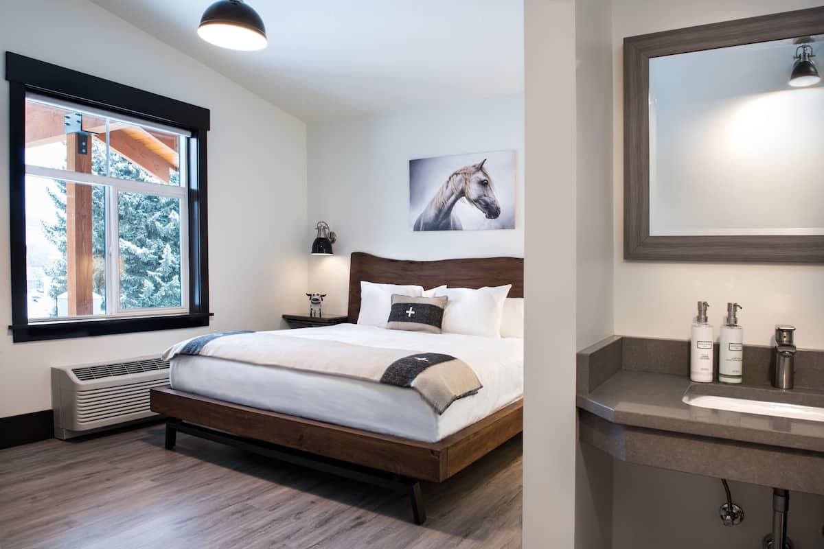 A bedroom with a large bed, a window with a view of trees, a mounted horse painting, and a bathroom with a sink and toiletries.