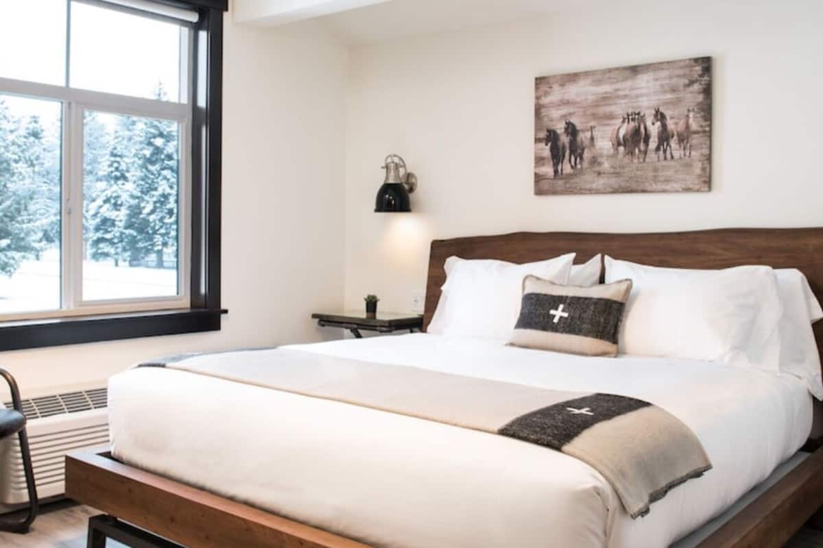 A bedroom with a wooden bed frame, white bedding, a wall-mounted lamp, and a painting of horses.