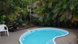 Une piscine ronde, bordée de blanc, entourée d’une végétation luxuriante et agrémentée d’une chaise en plastique blanche.