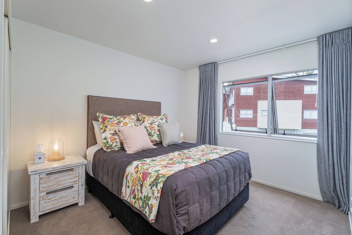 Une chambre à coucher avec un lit, une table de chevet, une fenêtre avec des rideaux et une vue sur un bâtiment à l’extérieur.