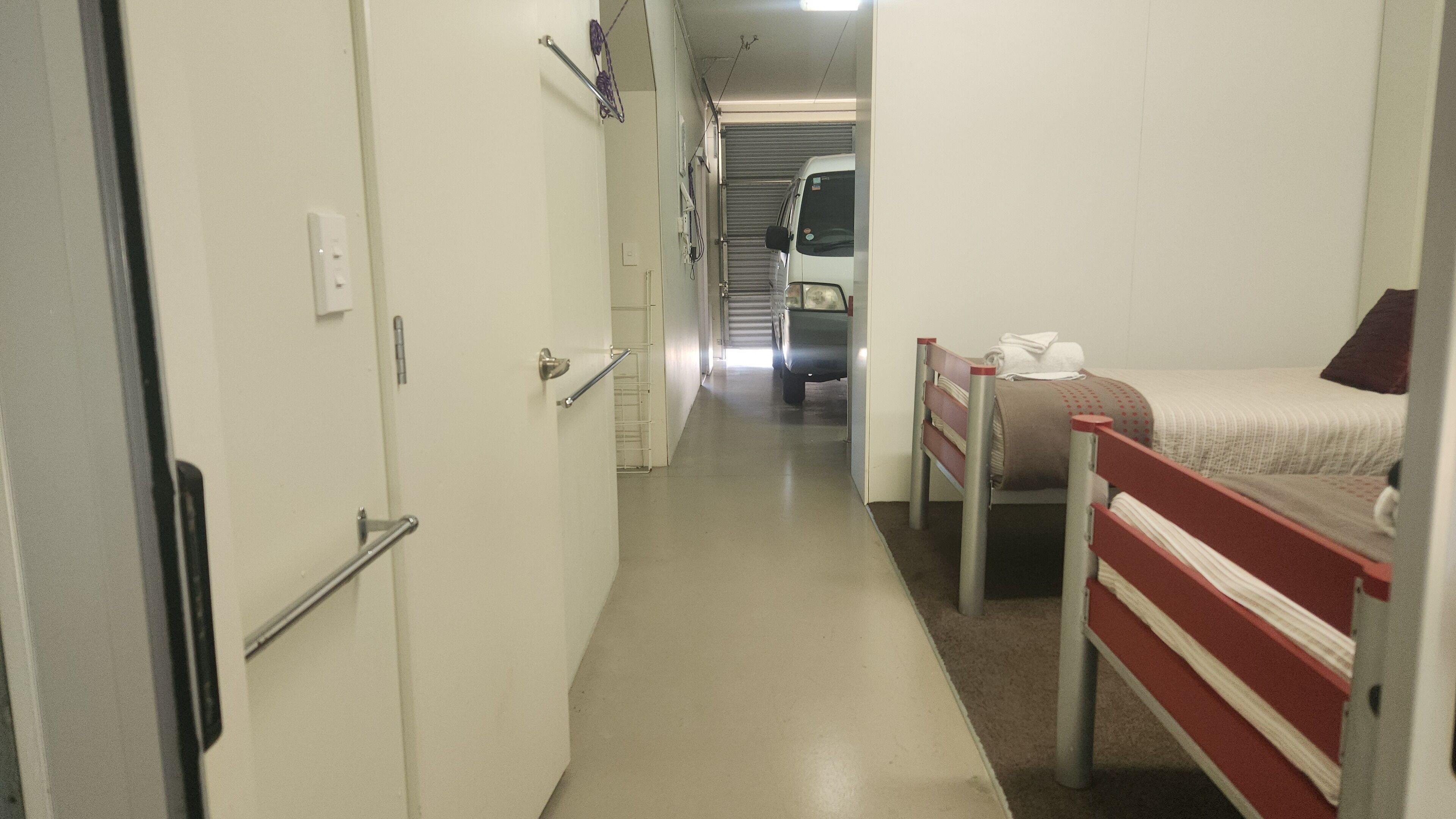 Um corredor estreito com uma cama à direita, uma porta à esquerda e uma van estacionada ao fundo.