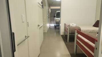 Um corredor estreito com uma cama à direita, uma porta à esquerda e uma van estacionada ao fundo.