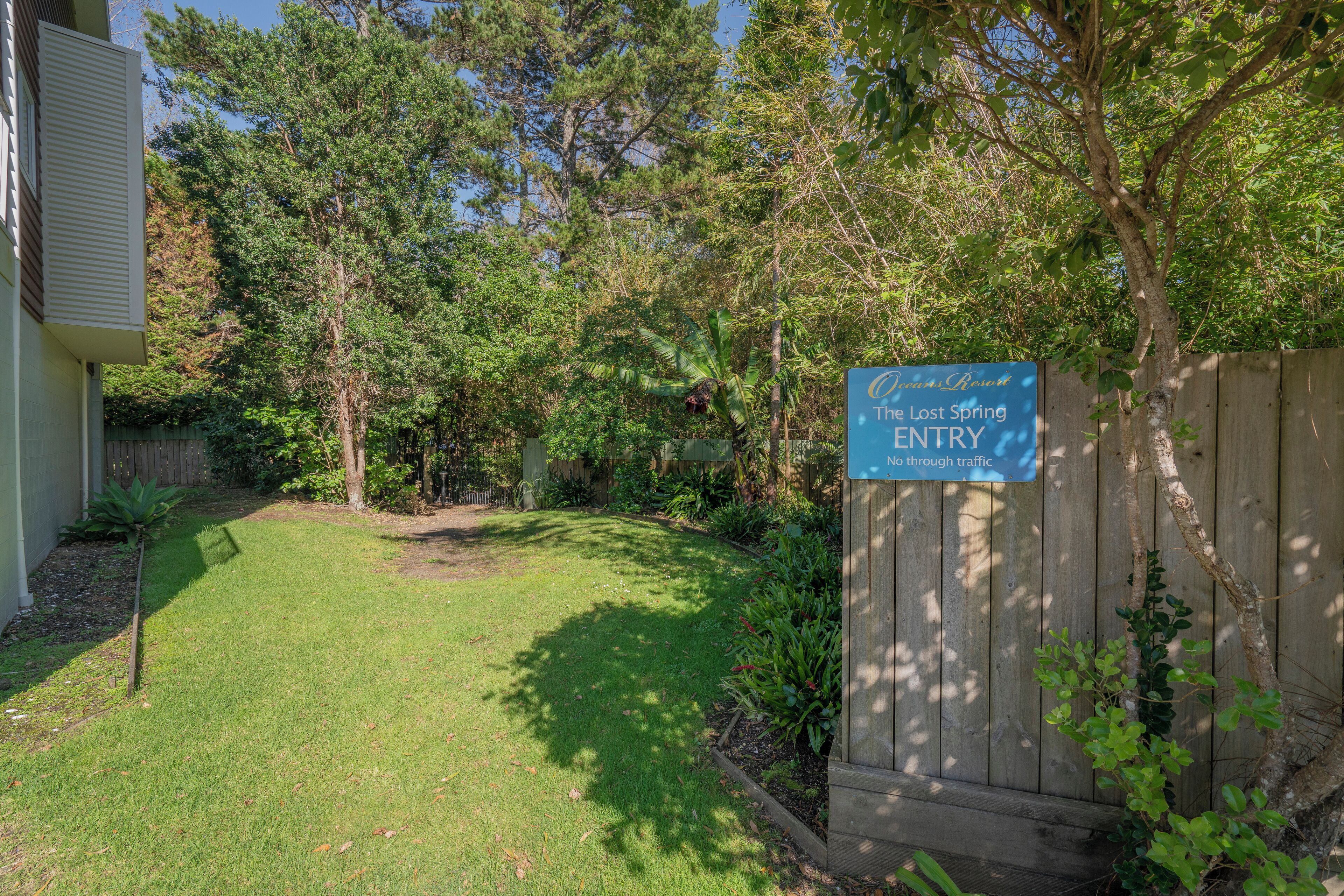A backyard with a wooden fence, a sign reading "The Lost Spring ENTRY No through traffic," and various trees and plants.