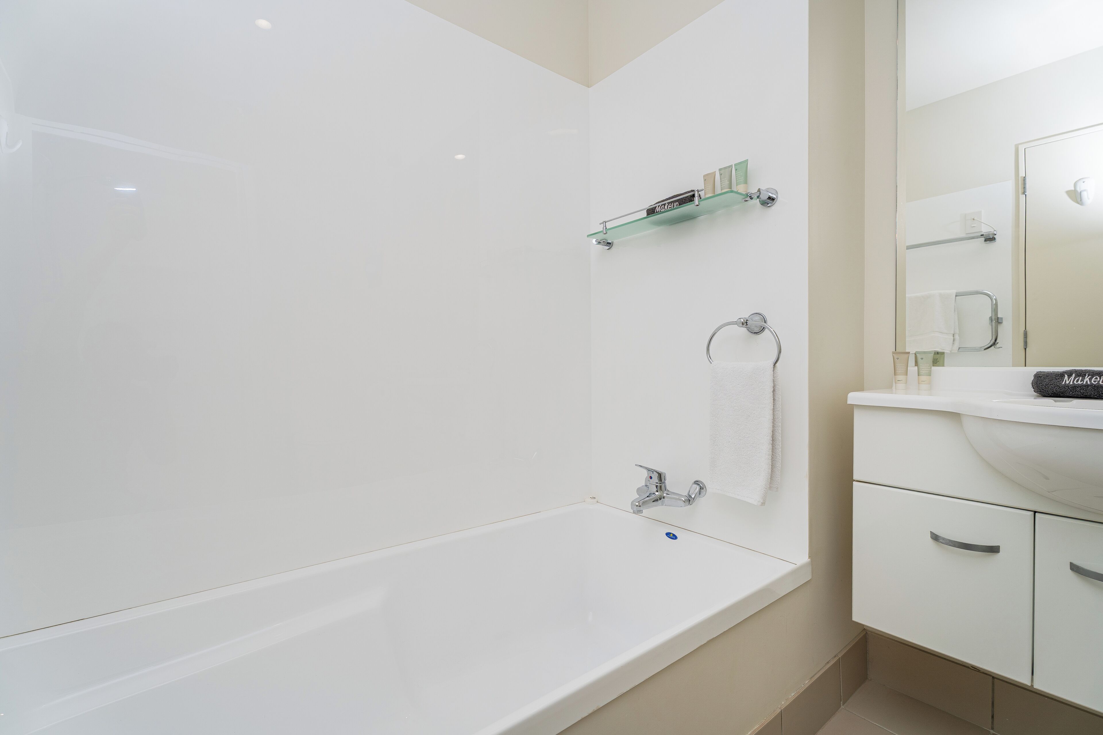 Un baño moderno con una bañera blanca, un estante de vidrio con artículos de aseo, un perchero y un tocador con espejo.