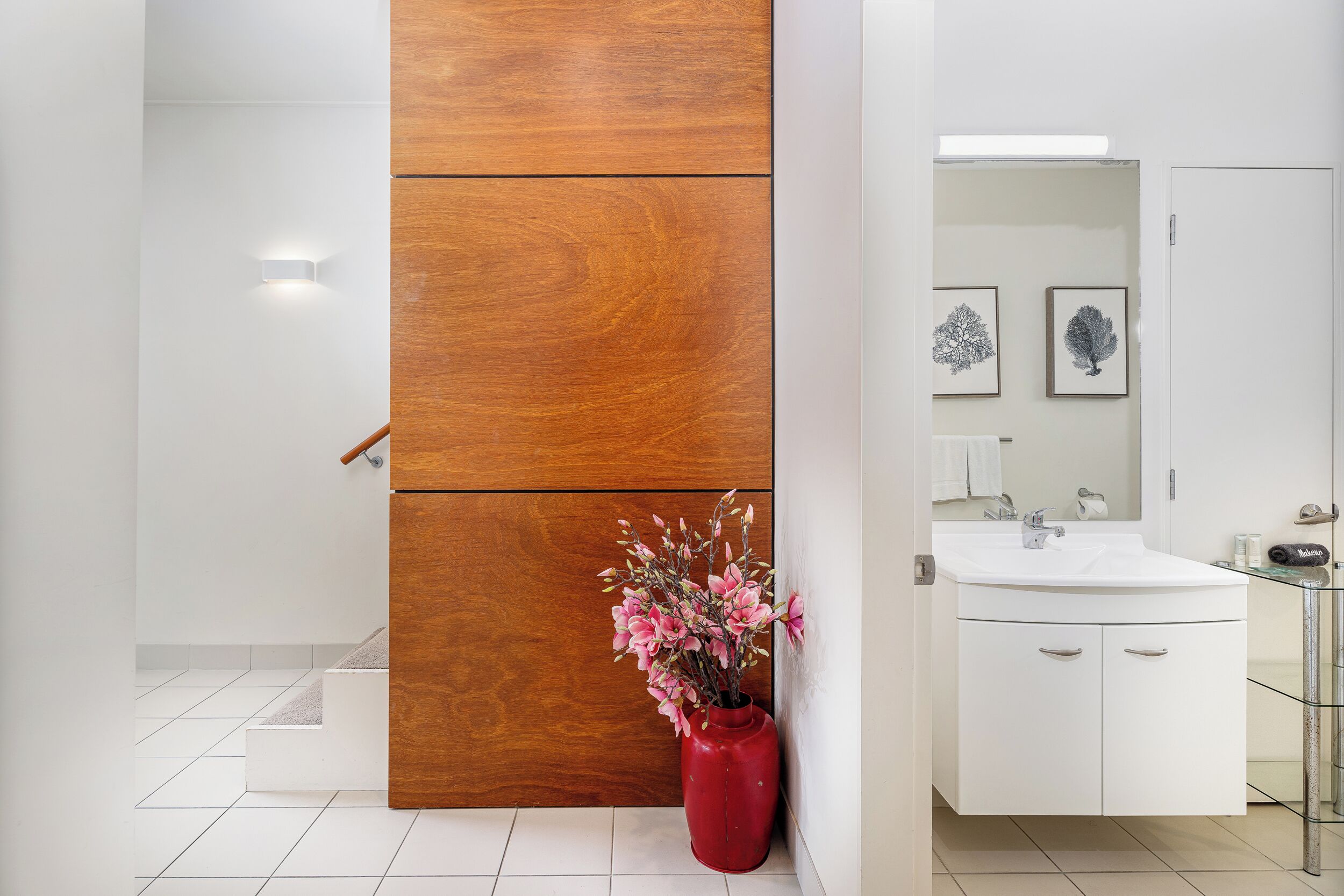 A modern bathroom with a wooden panel, a white sink, and a red vase with flowers.