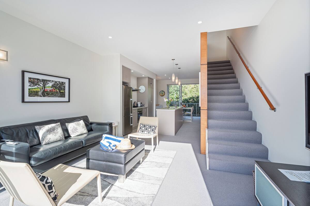 Un salon moderne avec un escalier gris, un canapé en cuir noir et un coin repas à l’arrière-plan.