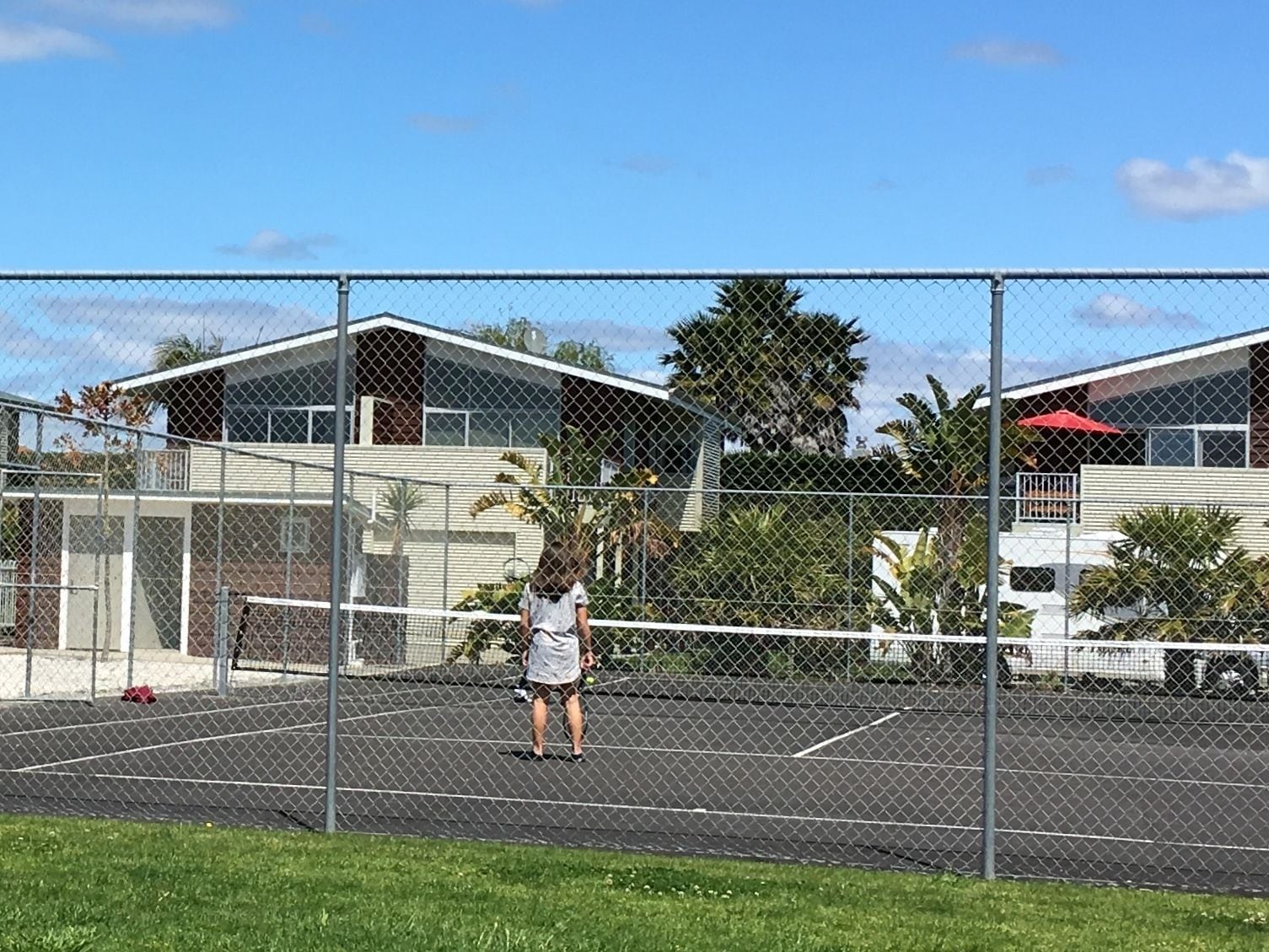 A fenced tennis court with a net, surrounded by buildings and greenery.