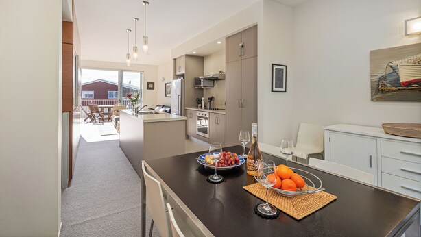 A modern kitchen with a central island, a dining area with a table and chairs, and a view of a house through a glass door. - Ocean Serenity Apartments Whitianga (Whitianga)