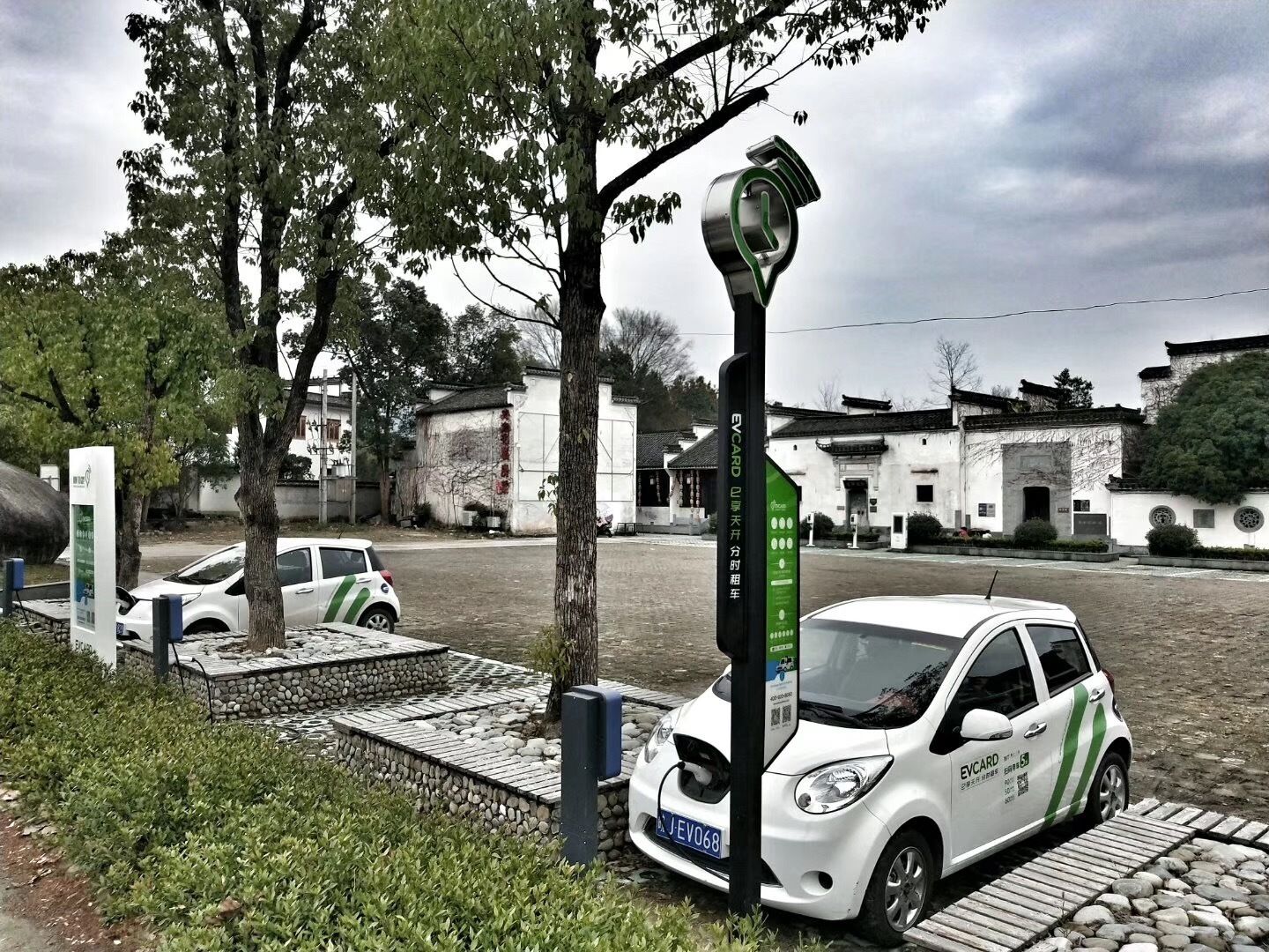 Une voiture électrique blanche est garée près d’une borne de recharge, avec des bâtiments à l’architecture traditionnelle en arrière-plan.