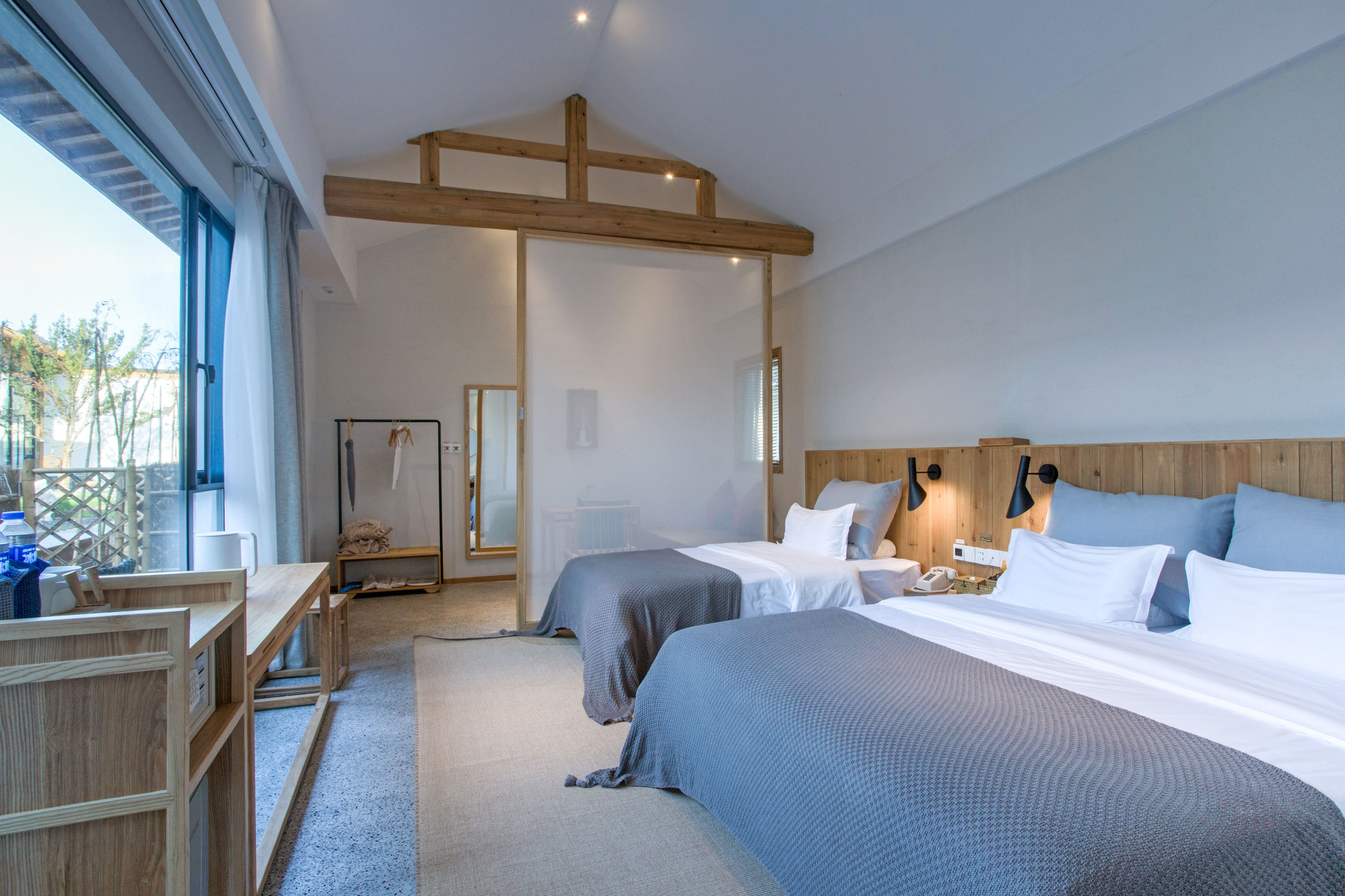 A modern hotel room with two beds, a wooden ceiling, a large window, and a small desk.