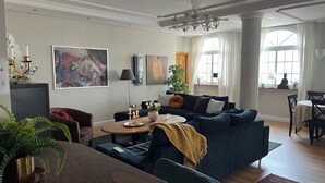 A modern living room with a dark blue sofa, a wooden dining table, and a chandelier. There are paintings on the walls and a large window with curtains. - Penthouse Duplex, Fuengirola (Los Pacos) (Fuengirola)