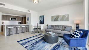Un salon moderne avec un canapé gris, un fauteuil bleu et une table basse. Il y a également un coin cuisine avec un bar et des tabourets.