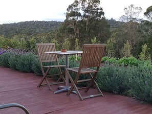 A wooden deck with two chairs and a table, surrounded by lush greenery and a forested hill in the background. - Lockleigh Park - Escape to the Country (Lebrina)