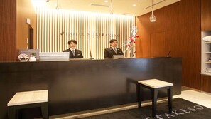 En hotellreception bemannad av två personer, en reception i trä och ett receptionsområde med en svart matta och en skylt med texten "Hotel Nuretsu".