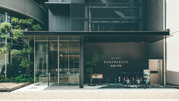 A modern hotel entrance with a glass facade, a sign reading "HOTEL KURETAKESO," and several bicycles parked outside. - Hotel Kuretakesou Hiroshima Otemachi (Hiroshima)