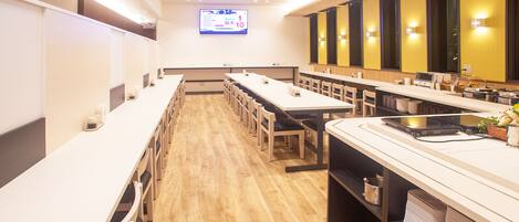 Una sala de conferencias moderna con largas mesas, sillas, una pantalla grande y una zona de cocina con electrodomésticos.