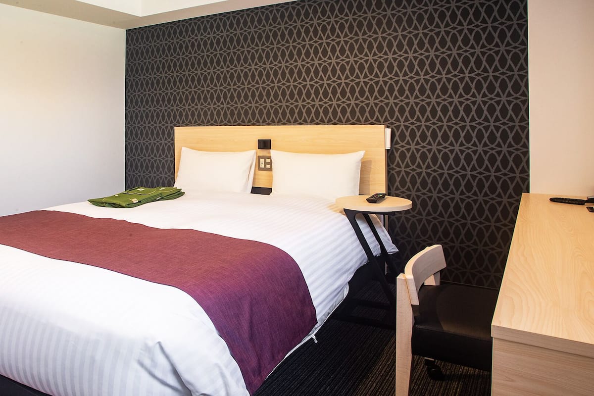A neatly made bed with a wooden headboard, a bedside table with a remote control, and a chair.