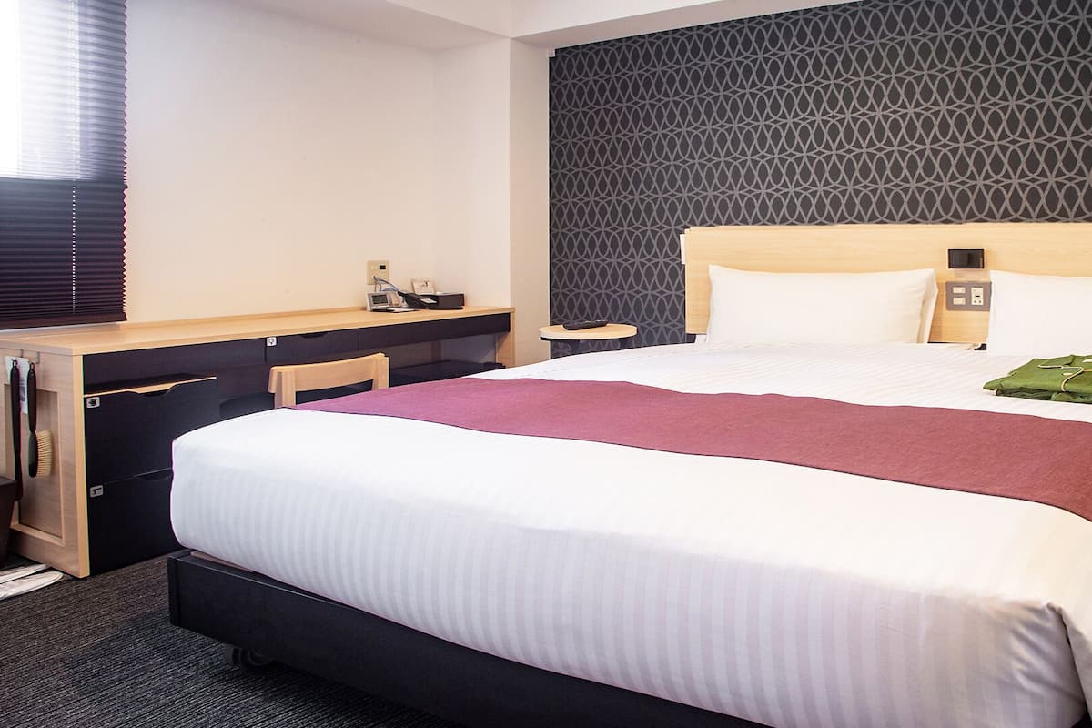 A hotel room with a large bed, a desk, a chair, a small table, and a patterned wall.
