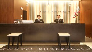 A hotel reception desk with two staff members. The reception area has a wooden backdrop with the hotel's name. There are two small tables in front of the desk. - Hotel Kuretakesou Hiroshima Otemachi (Hiroshima)